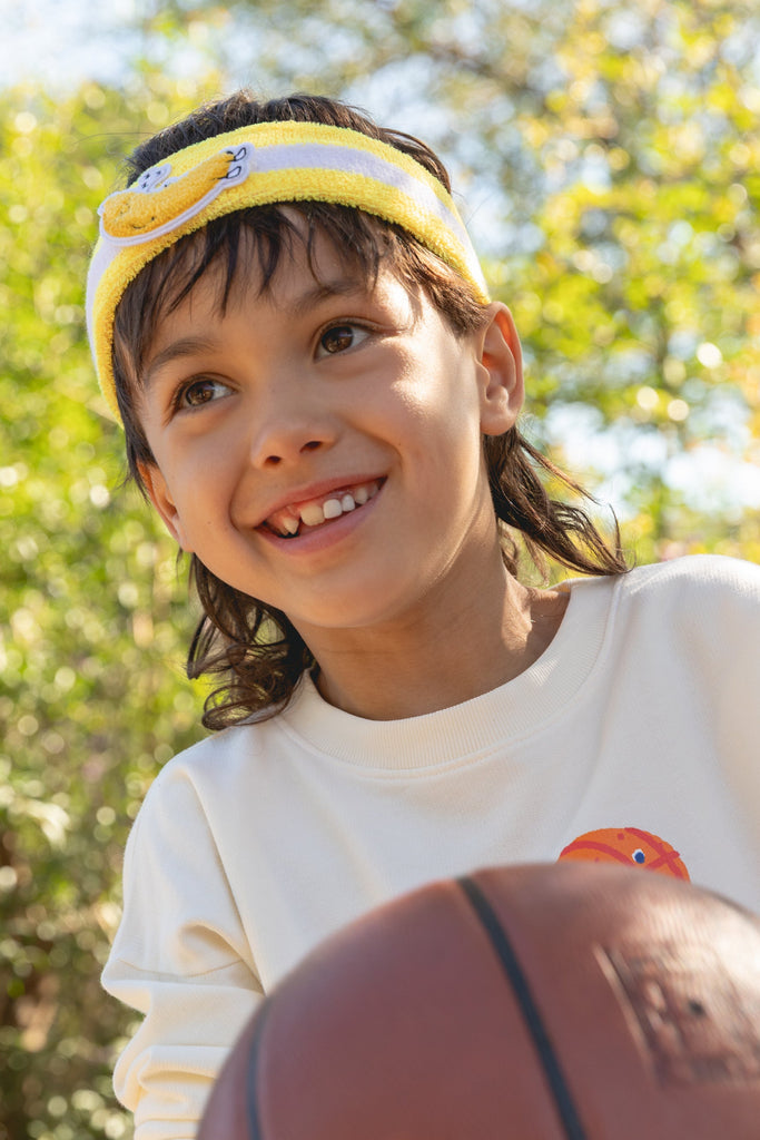 Child wearing a Dodo Banana headband and holding a basketball outdoors with greenery in the background