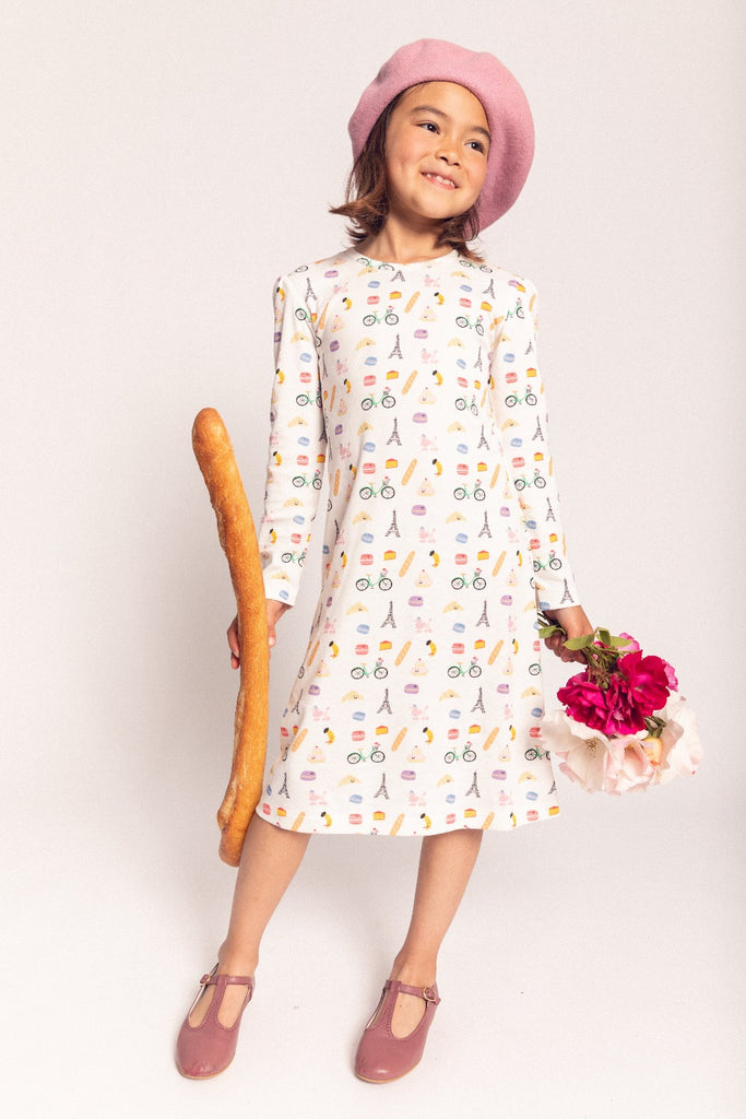 A little girl smiling while wearing a hat, holding a baguette and flowers, while wearing a Frenchie Long Sleeve night gown from Dodo Banana